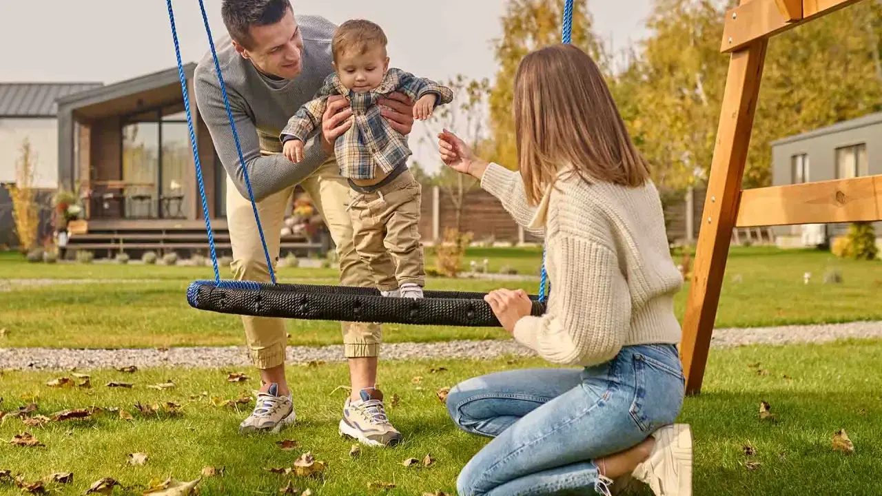 Radosne zabawy w ogrodzie: tata podtrzymuje dziecko na huśtawce, mama zachęca do zabawy.