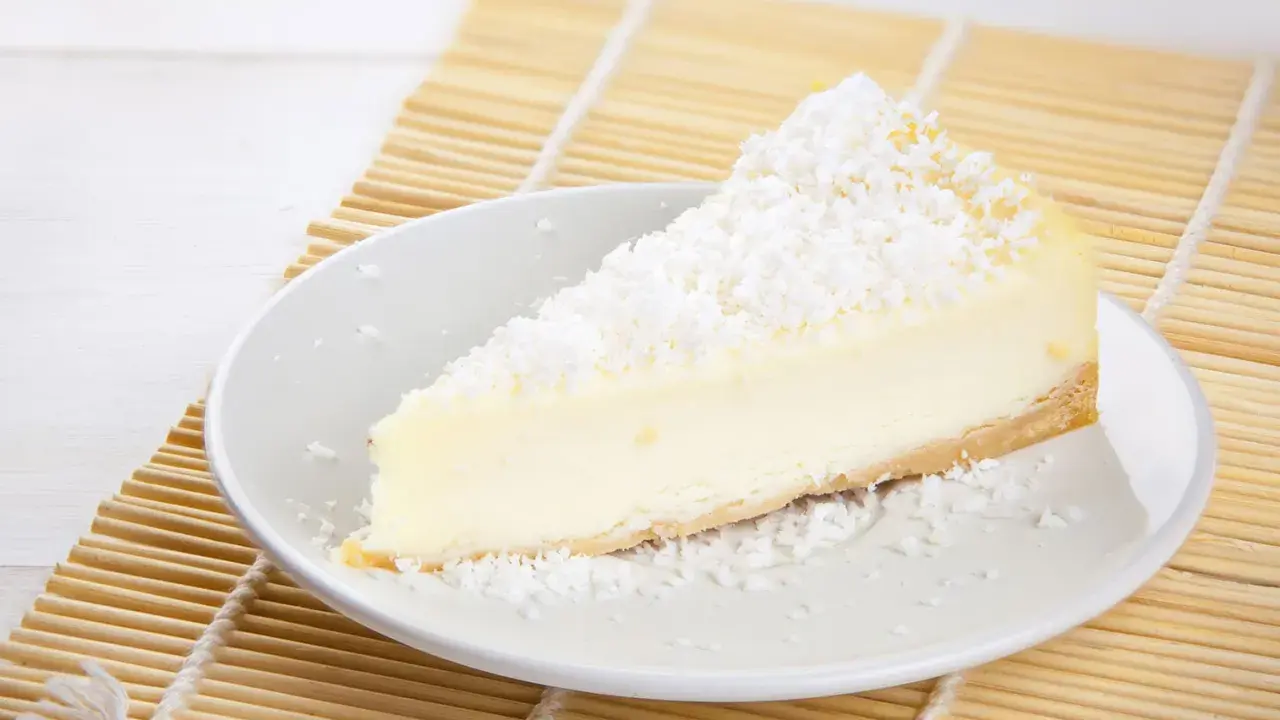 Une part de tarte noix de coco, crémeuse et généreusement saupoudrée de noix de coco râpée, repose sur une assiette blanche, prête à être dégustée.