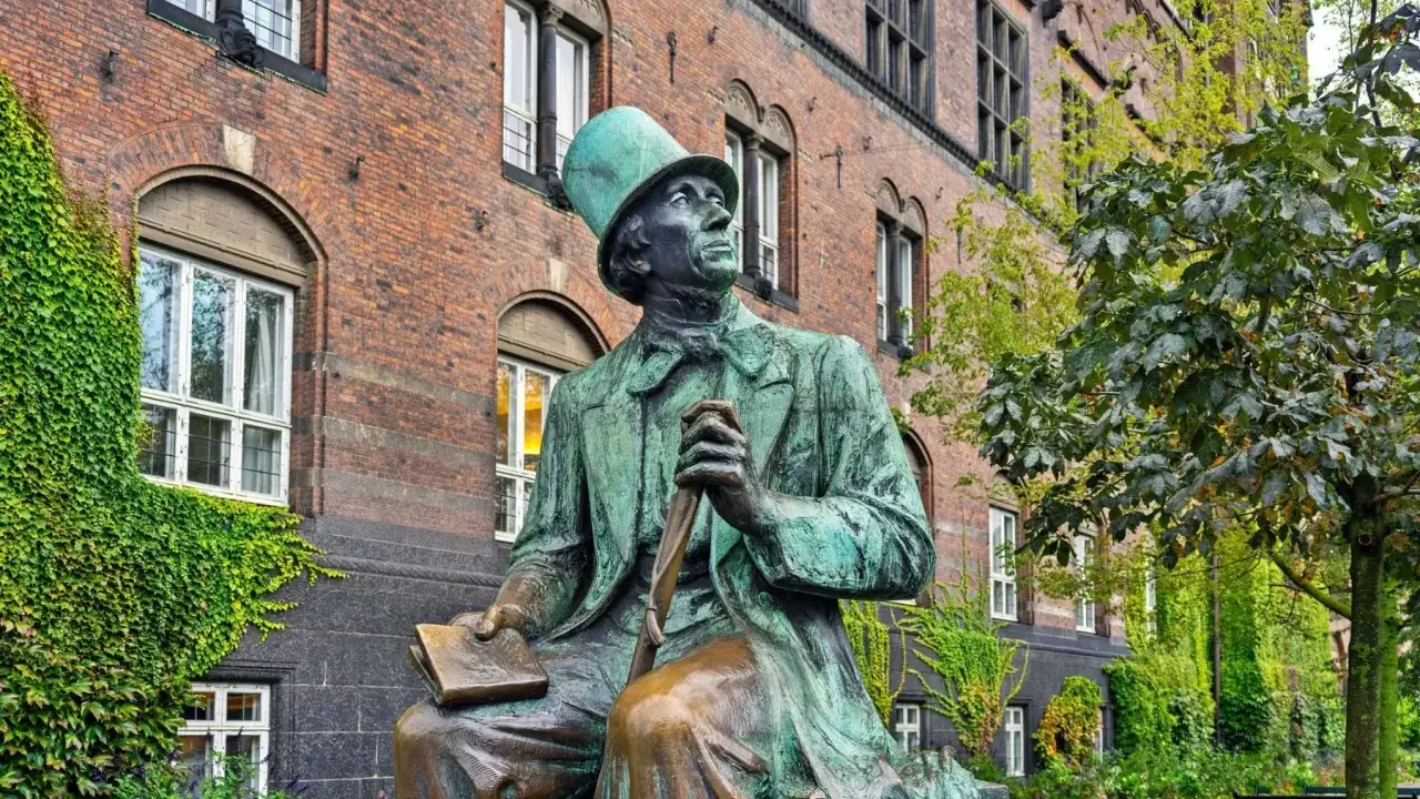 Pomnik Jana Christiana Andersena w Kopenhadze, siedzącego z książką i laską, na tle ceglanego budynku porośniętego bluszczem.