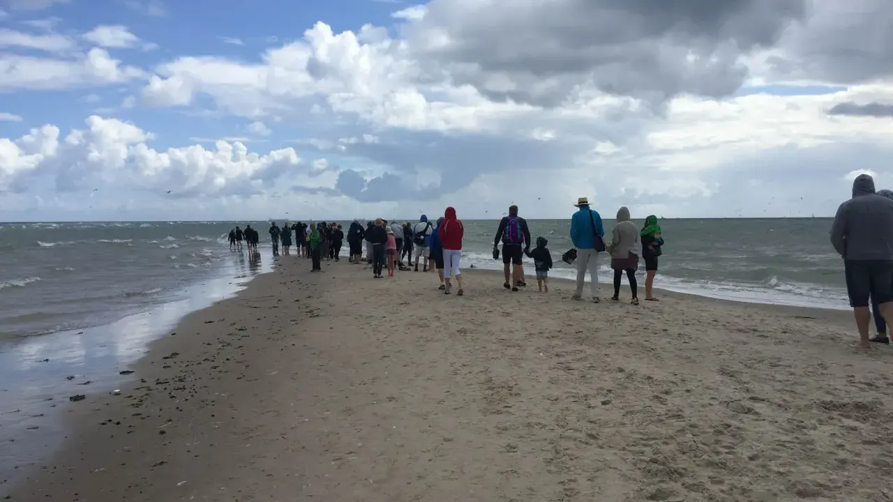Folk promenerar längs en sandremsa vid vattnet norr om Skagen. Molnig himmel och hav.