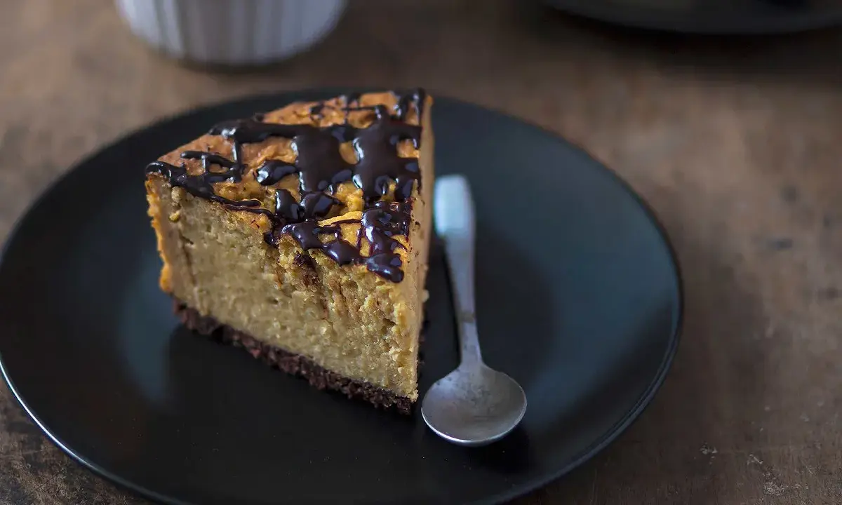 Tranche de cheesecake sans gluten sans lactose, nappée de chocolat noir, sur une assiette noire avec une cuillère.