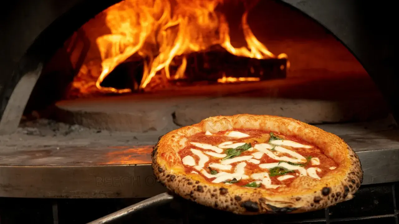 En perfekt italiensk pizza med mozzarella och basilika tas ur en vedeldad ugn med flammande eld.