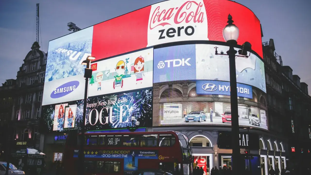 Wielkie ekrany reklamowe na placu, prezentujące różne rodzaje reklamy: Coca-Cola Zero, Samsung, Vogue, Hyundai, TDK. Widać też autobus i ludzi.