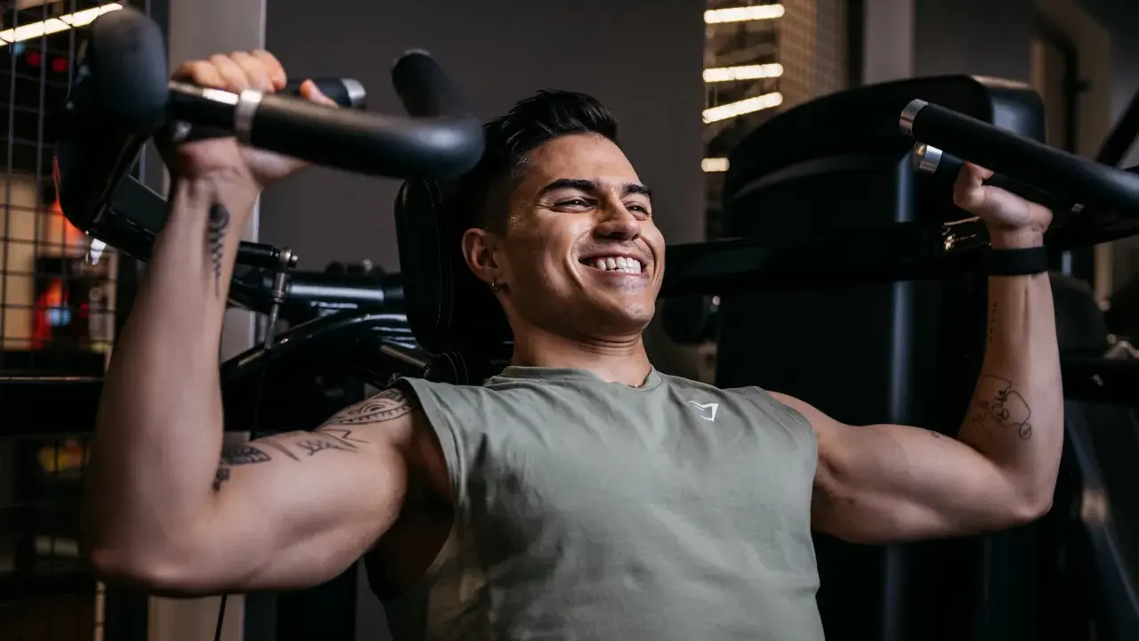 Ein Mann trainiert im Fitnessstudio, um seinen Brustmuskel aufzubauen. Er lächelt zufrieden, während er die Gewichte hebt.