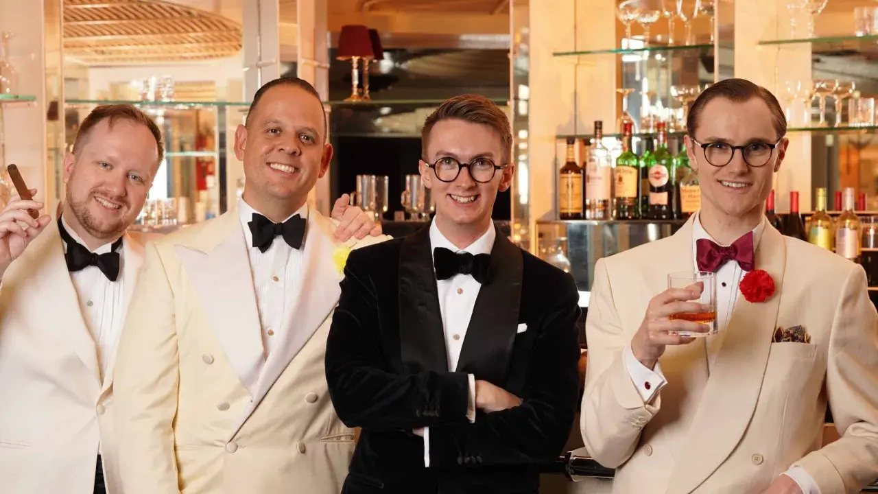 Four men in stylish suits and bow ties, embodying creative black tie, pose at a bar. One holds a cigar, another a drink.