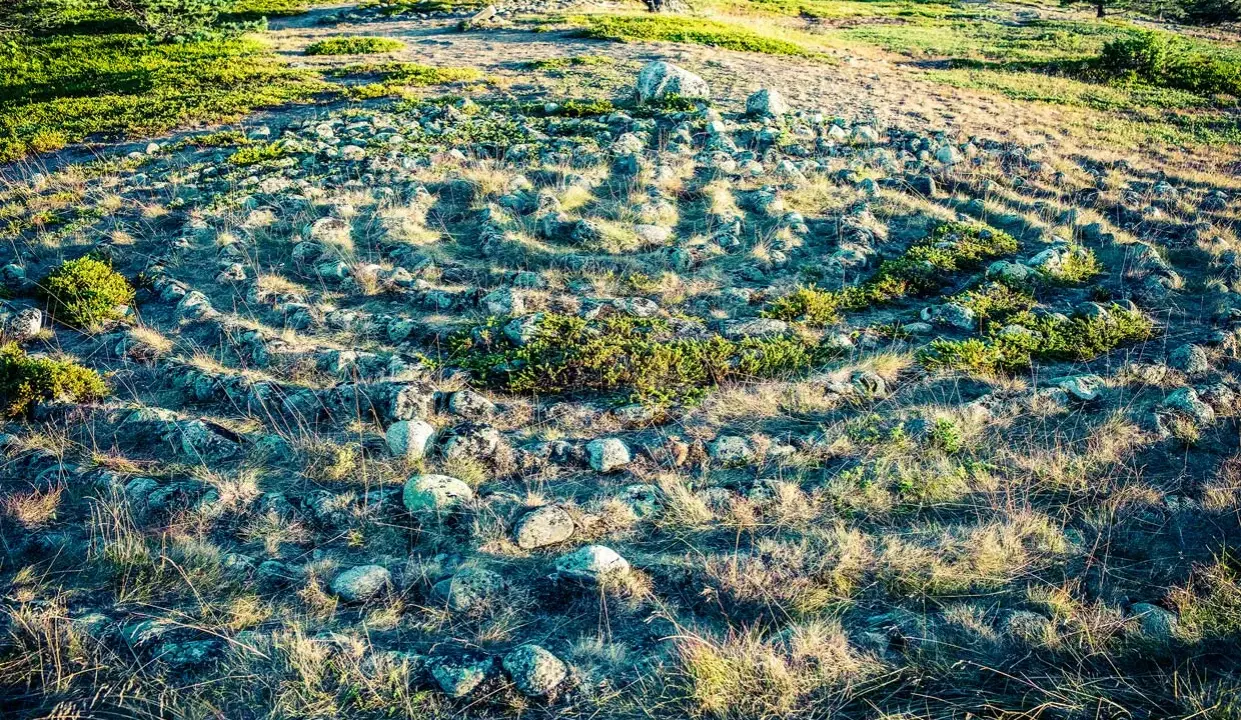 Stenlagd labyrint i Sverige, omgiven av gräs och buskar. Solen kastar långa skuggor över den uråldriga stensättningen.