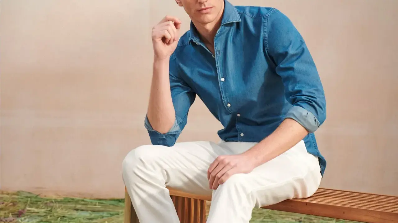 A man poses in a blue denim shirt and crisp white jeans, looking relaxed on a wooden bench.