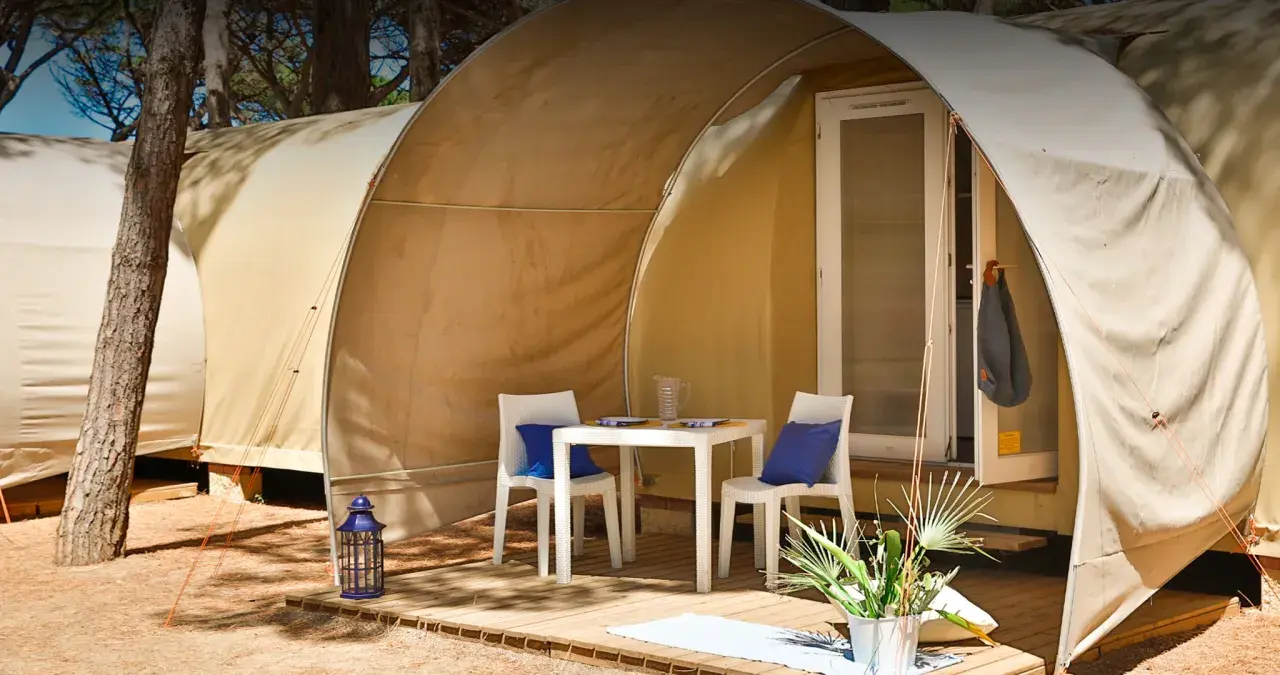 Tavolino e sedie con cuscini blu davanti a una tenda del **Gitavillage Il Gabbiano**. Lanterna blu e piante sul deck.