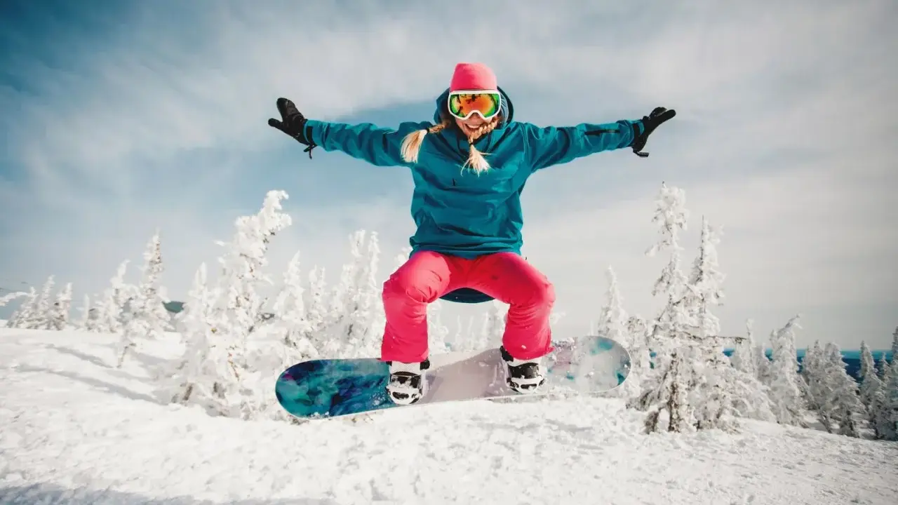 Dziewczyna w różowej czapce i goglach skacze na snowboardzie na tle zaśnieżonych drzew. To jest właśnie snowboard!