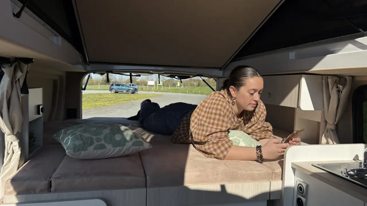 Une jeune femme se détend sur le lit de son camping-car j80 interieur, regardant son téléphone. Dehors, le soleil brille.