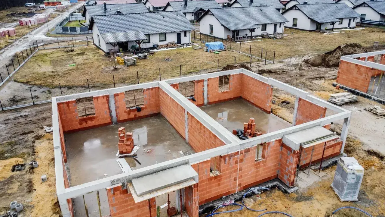 Budowa osiedla domów jednorodzinnych. Fundamenty i ściany parteru wznoszone z cegły, w tle gotowe domy. To początek drogi, jak zostać deweloperem.