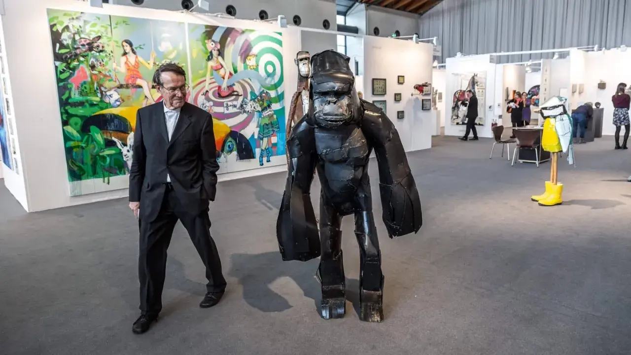Mann in Anzug neben Gorilla-Skulptur auf Kunstmesse. Bunte Gemälde im Hintergrund zeigen aktuelle Kunst.