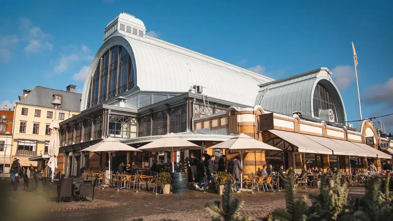 Människor sitter vid bord utomhus framför den imponerande **stora saluhallen** under en klarblå himmel.