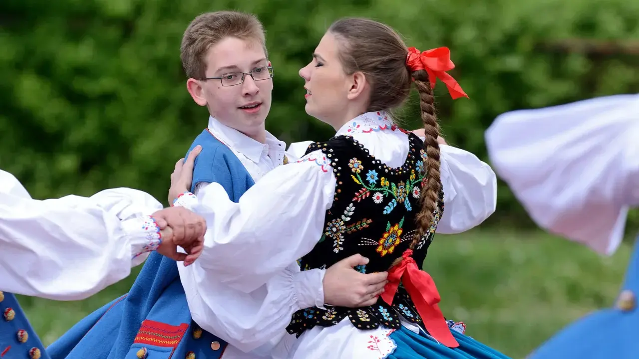 Młoda para w strojach ludowych tańczy mazura. Chłopiec w niebieskiej kamizelce, dziewczyna w haftowanej kamizelce i warkoczu z czerwoną wstążką.