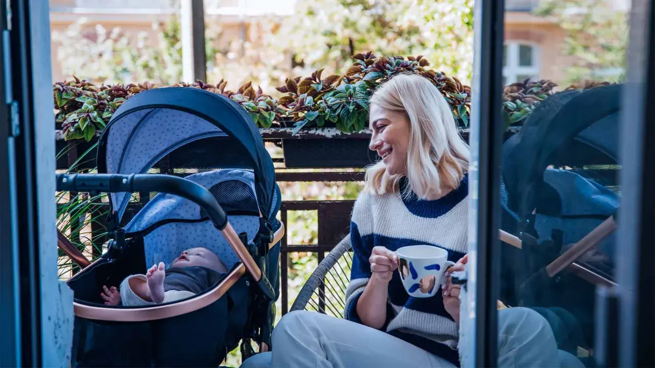 Uśmiechnięta mama z kawą na balkonie, obok wózek z dobrą amortyzacją i śpiące dziecko.