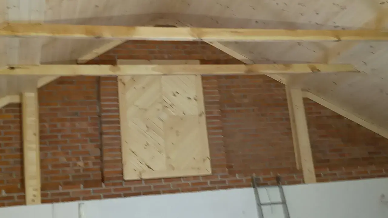 Drewniana konstrukcja dachu nad ceglaną ścianą, z ozdobnym panelem. Stara więźba dachowa czeka na renowację.