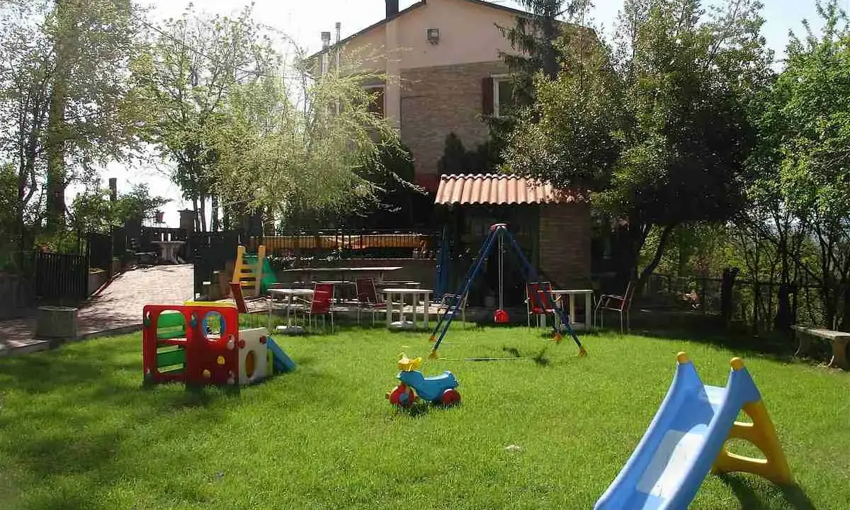Un ristorante con fattoria offre un'area giochi per bambini con scivolo, altalena e giochi colorati, immerso nel verde.