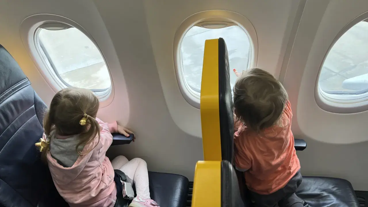 Dzieci patrzą przez okno samolotu Wizz Air. Dziewczynka w różowej bluzie i chłopiec w pomarańczowej koszulce, siedzą obok siebie.