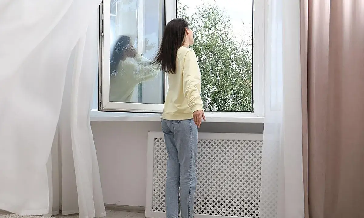 Mujer abre ventana para ventilar la casa en invierno. Reflejo en el cristal muestra a otra persona.