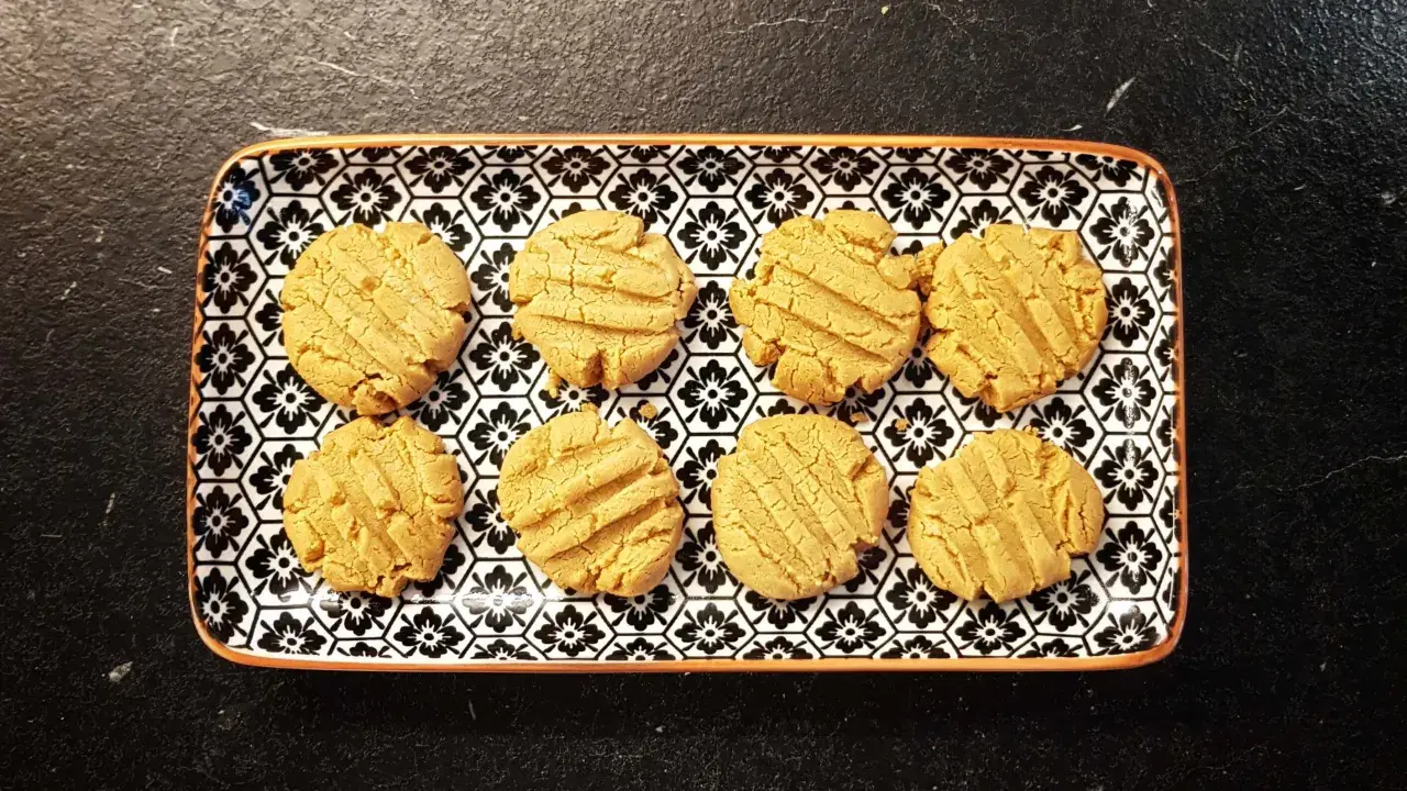 Acht zuckerfreie Erdnussbutter Haferflocken Kekse liegen auf einem rechteckigen Teller mit schwarzem Blumenmuster.