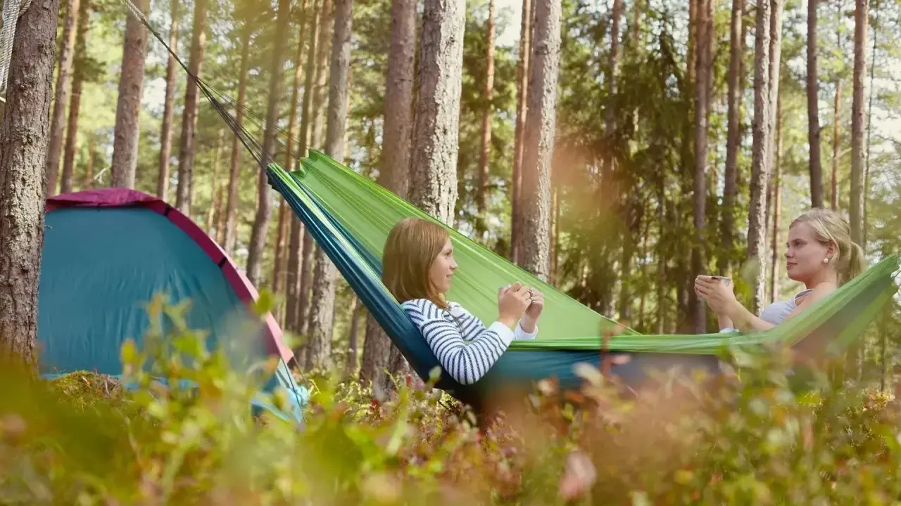 Två kvinnor njuter av en lugn stund i en hängmatta i skogen, med ett tält i närheten. En perfekt bild av campa Sverige.