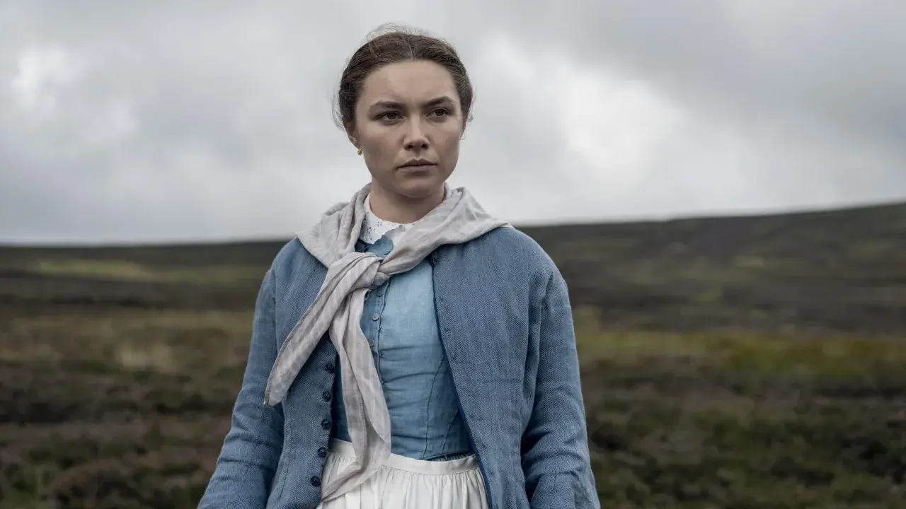 Florence Pugh i en blå klänning och jacka, med en scarf runt halsen, står i ett öppet landskap. En scen från filmer med Florence Pugh.