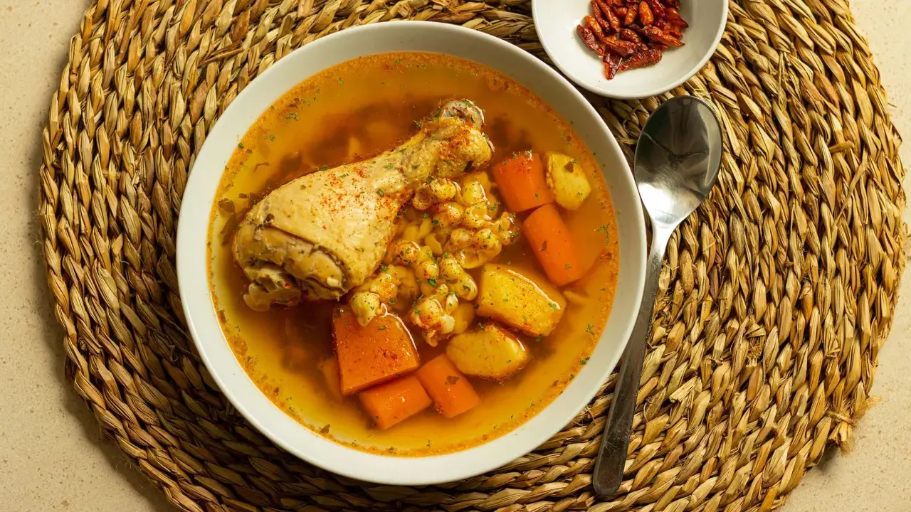Un confortante brodo di pollo con pezzi di pollo, carote e patate, servito su una tovaglietta di paglia.
