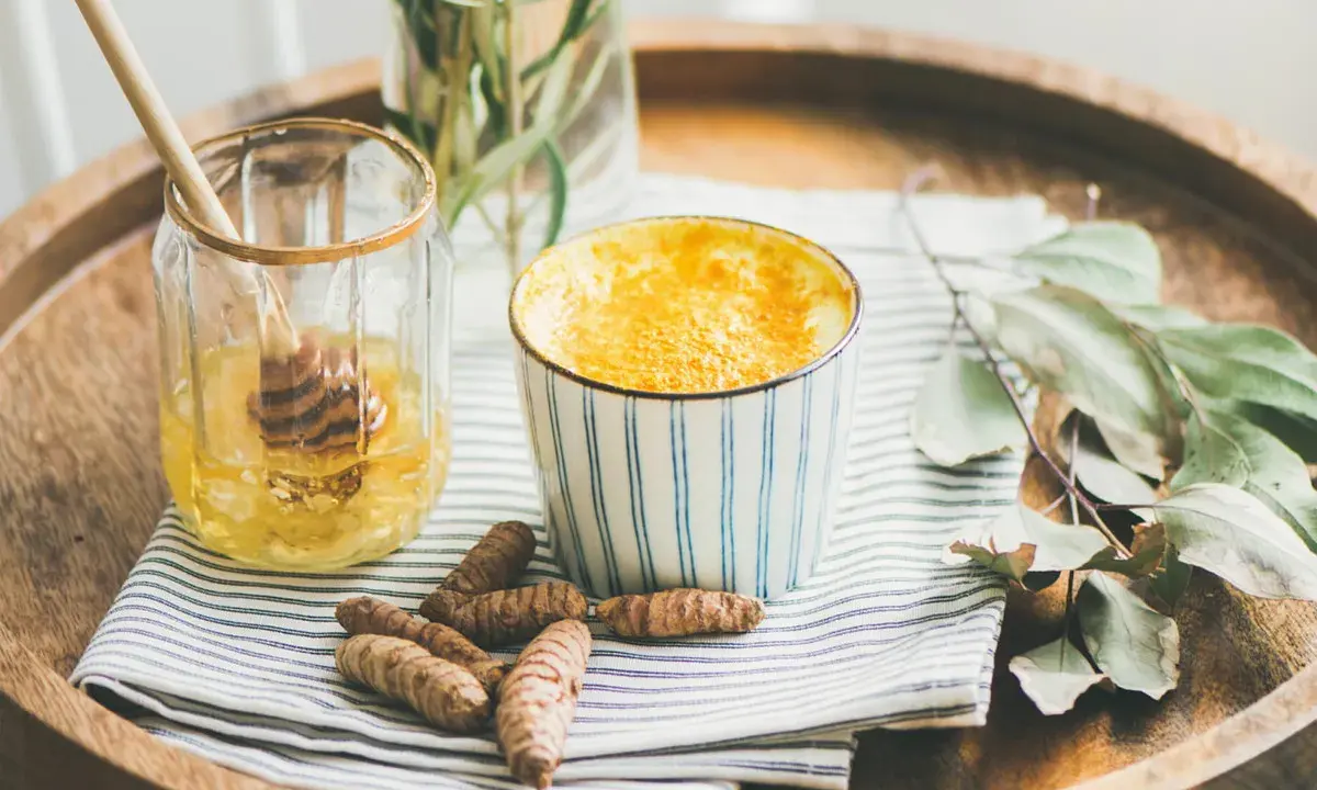 Aromaticzne złote mleko w kubeczku, obok słoiczek z miodem i korzenie kurkumy. Idealne na chłodne wieczory.