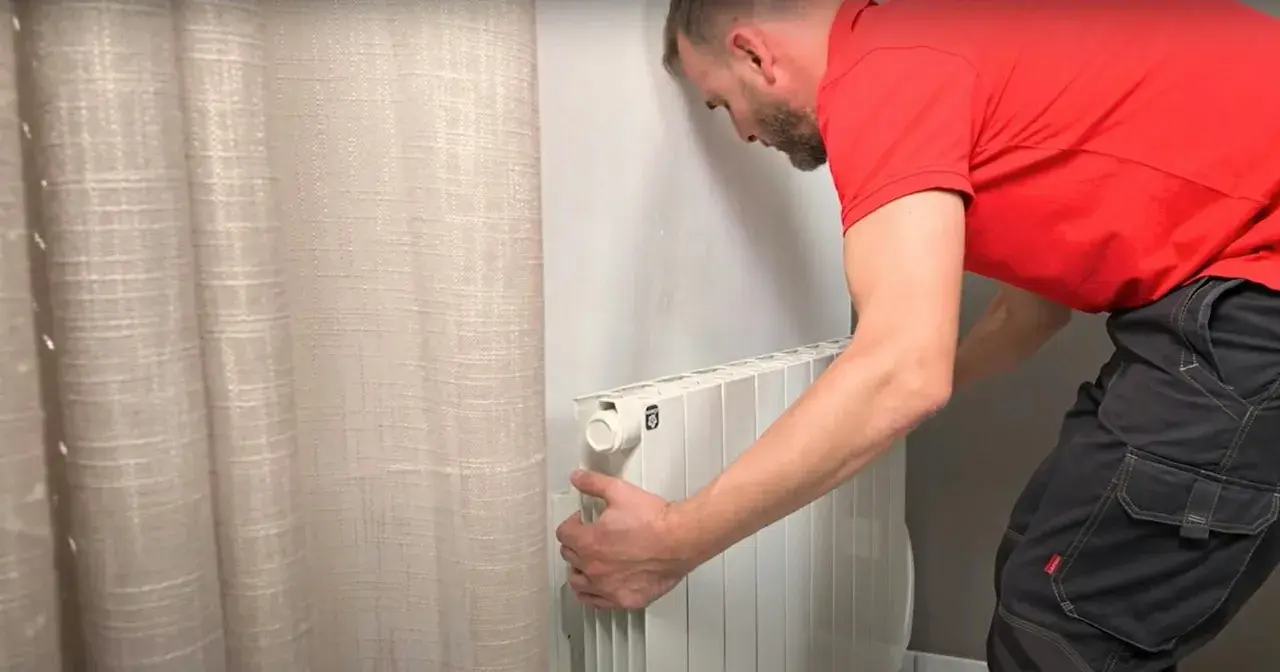 Un homme en t-shirt rouge et pantalon de travail ajuste un radiateur électrique blanc, prêt pour son branchement.