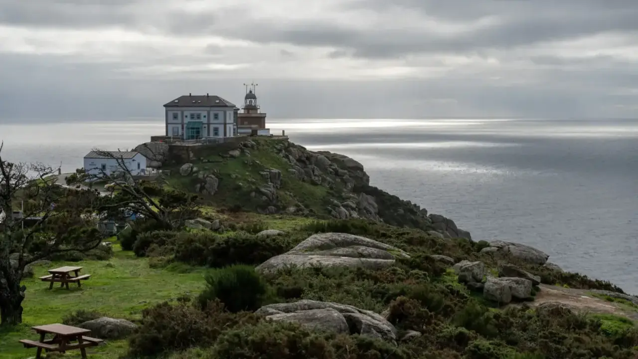 Leuchtturm und Gebäude auf felsiger Küste bei Finisterre, Spanien. Grauer Himmel über dem Meer.
