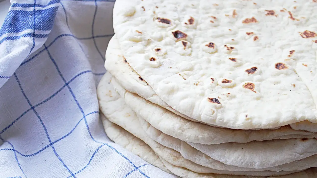En hög med nybakade tortillabröd, perfekta för ditt nästa tortilla recept. Bredvid ligger en blåvitrutig kökshandduk.