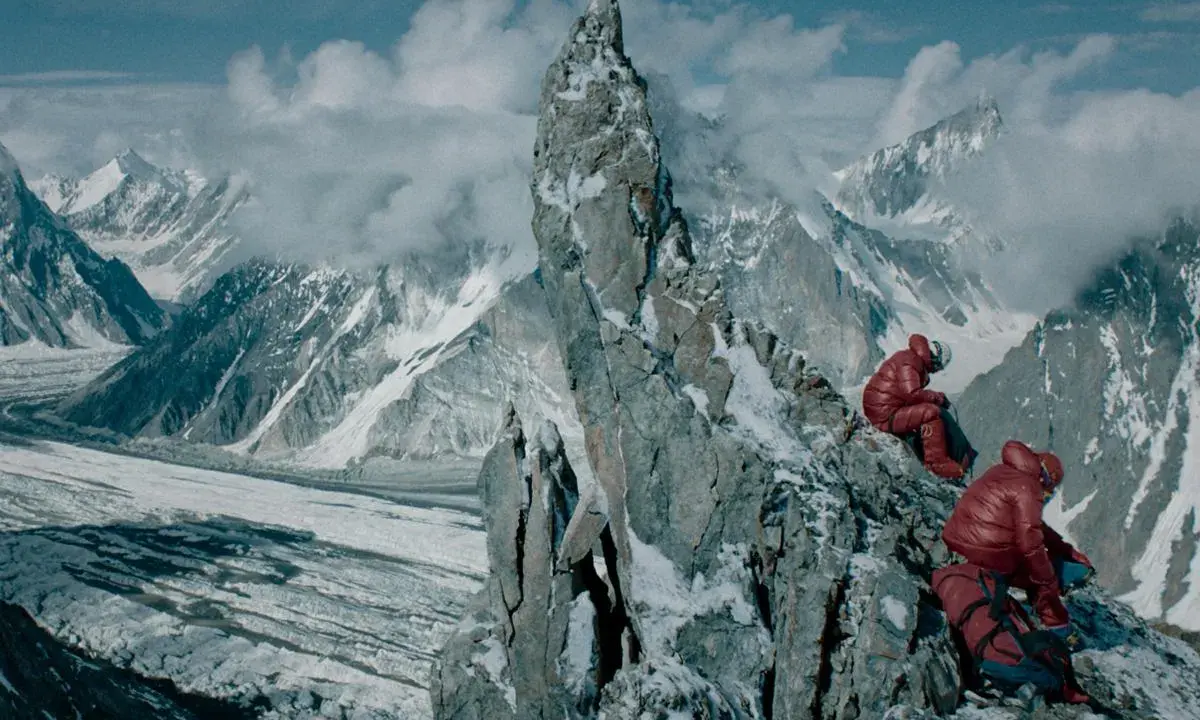 Dwóch alpinistów w czerwonych kurtkach odpoczywa na skalistej grani, podziwiając rozległy widok na lodowiec i ośnieżone szczyty. Broad peak góra w oddali.