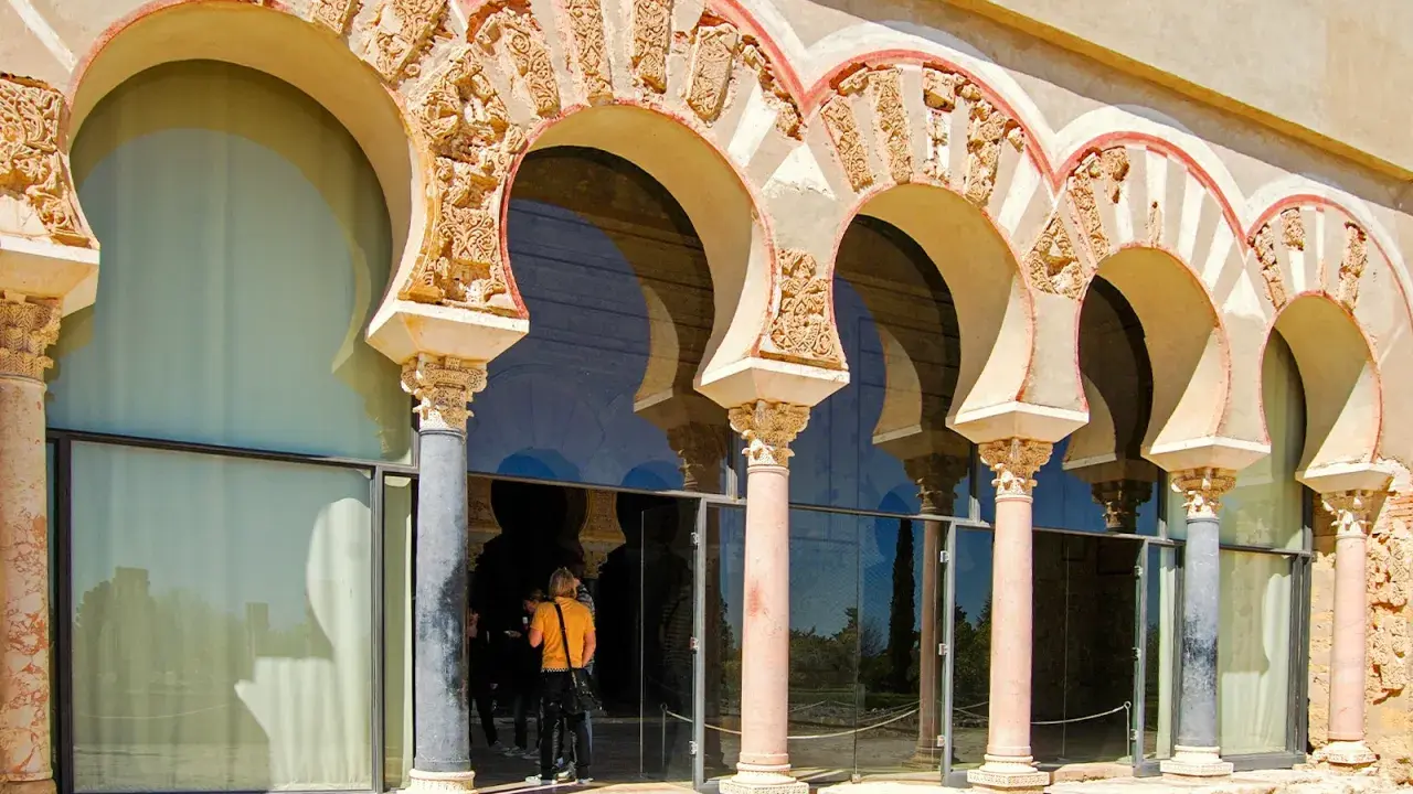 Verzierte Bögen und Säulen der Medina Azahara spiegeln sich in Glas. Besucher bestaunen die historische Architektur.