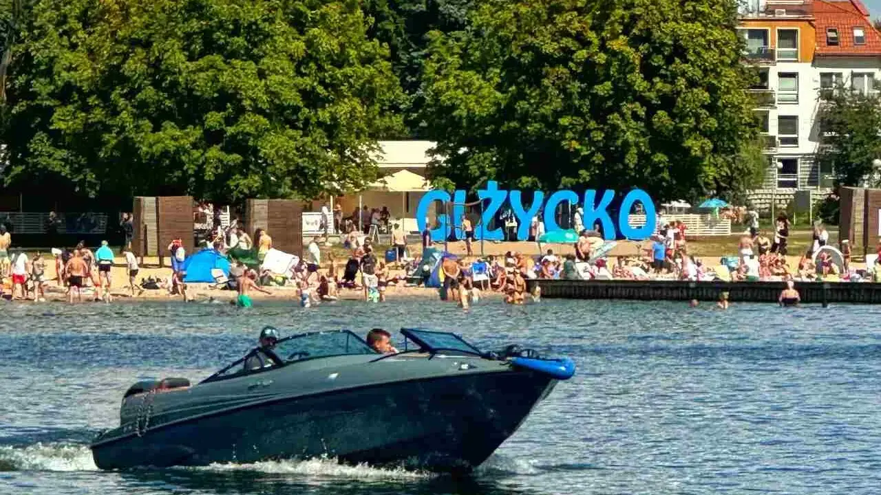 Motorówka sunie po wodzie, w tle plaża pełna ludzi i wielki napis "GIŻYCKO". Idealny dzień na Mazurach.