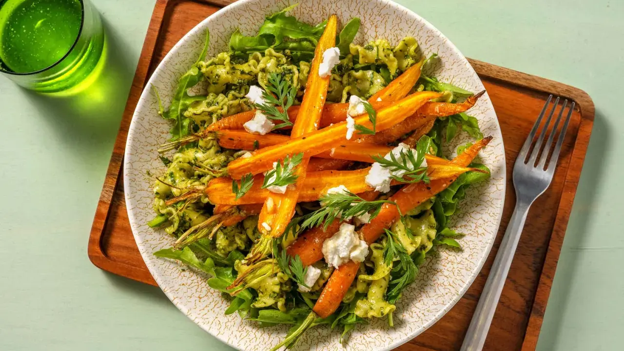 Un plat de pâtes vertes garni de carottes rôties et de fromage frais, avec des fanes de carottes pour la décoration.