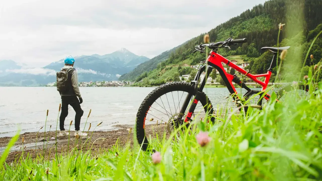 En person med hjälm och ryggsäck står vid en sjö med en mountainbike framför sig. Berg och en stad syns i bakgrunden. Perfekt för att cykla MTB.