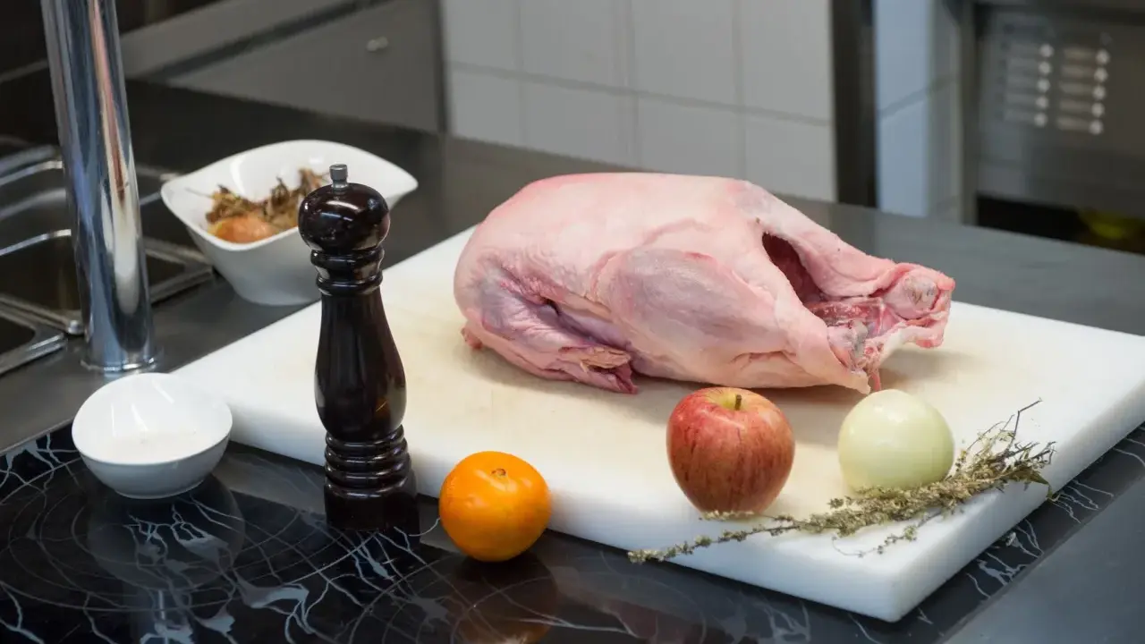 Rohe Gans auf einem Schneidebrett mit Apfel, Zwiebel, Orange, Gewürzen und Pfeffermühle. Die Frage "wie lange dauert auftauen" beschäftigt jeden Koch.