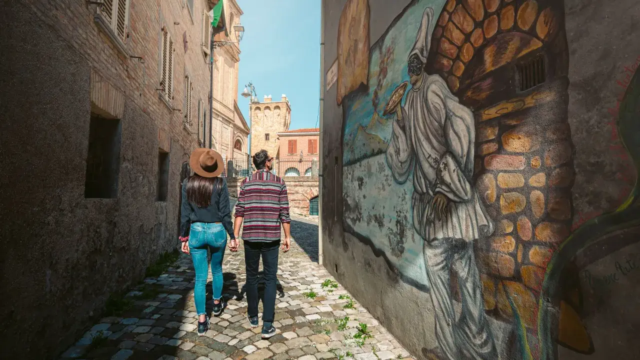 Coppia passeggia in un vicolo di Saludecio, ammirando un murale. Scopri cosa vedere in questo borgo incantevole.