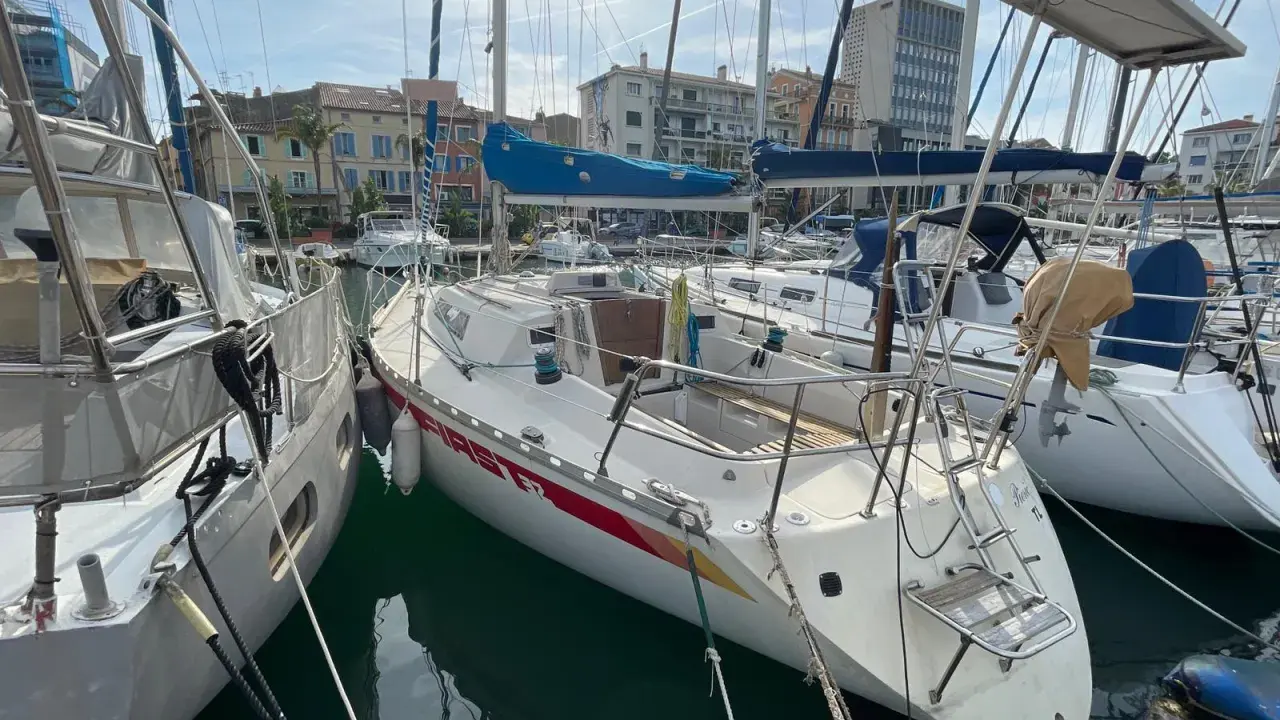 Un voilier First 32, amarré dans un port, avec d'autres bateaux et des bâtiments en arrière-plan.