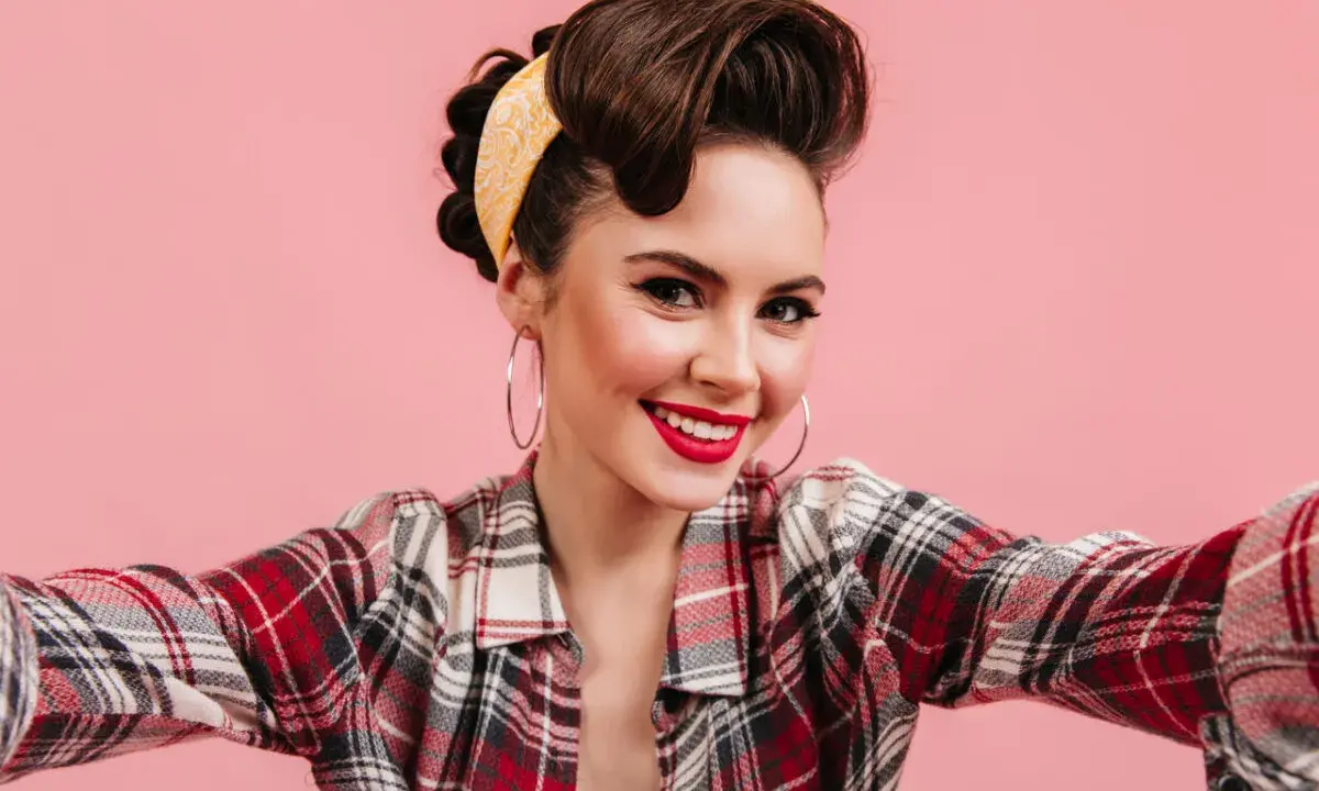 Uśmiechnięta dziewczyna w stylu pin up, z burzą brązowych włosów upiętych w kok i żółtą bandanką, w kraciastą koszulę.