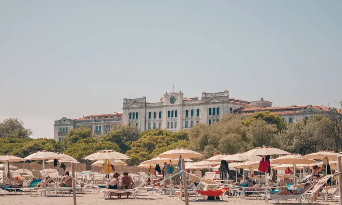 Entspannte Menschen liegen unter Sonnenschirmen am Lido di Venezia Beach. Im Hintergrund thront ein imposantes Gebäude vor üppigem Grün.