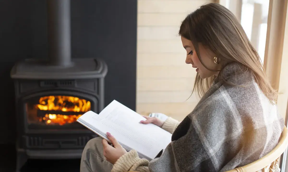 Une femme lit un livre près d'un poêle à granulés sans électricité. Avis : chaleur réconfortante et ambiance cosy.