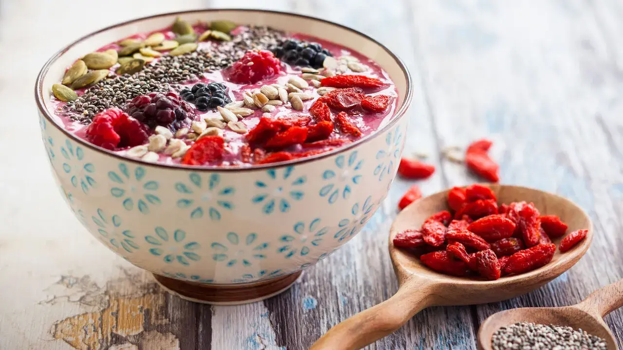 Miseczka smoothie z owocami, nasionami chia, pestkami dyni i słonecznika. Obok łyżka z jagodami goji. Nasiona chia jak jeść? Dodaj do smoothie!