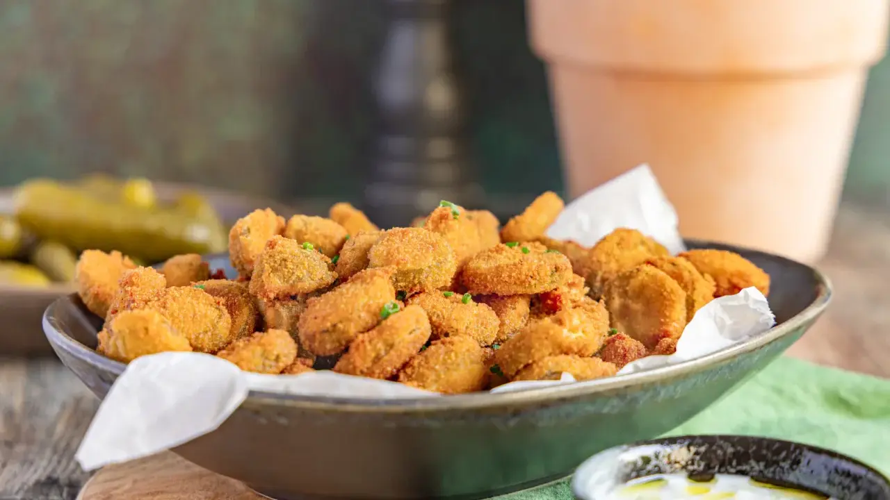 Knusprig frittierte Gurkenscheiben, **fried pickles**, in einer Schale, daneben ein Dip und ganze Gewürzgurken.