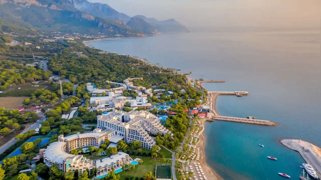 Widok z lotu ptaka na Rixos Sungate, luksusowy kurort nad turkusowym morzem, otoczony zielenią i górami.