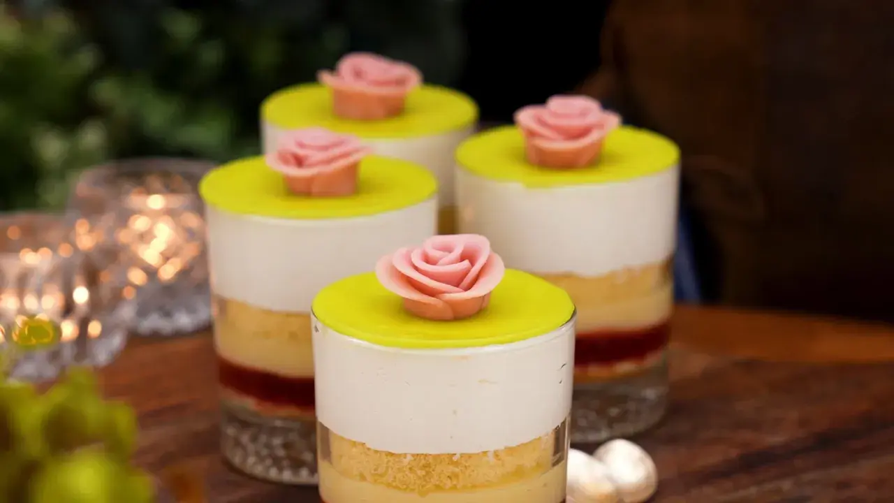 Fyra små prinsesstårta i glas, dekorerade med rosa marsipanrosor och gul marsipanlock. Vackert uppdukat med ljus i bakgrunden.