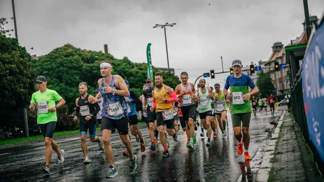 Uczestnicy maratonu biegną w deszczu po mokrej nawierzchni. Widać ich skupienie i determinację mimo niesprzyjającej pogody.
