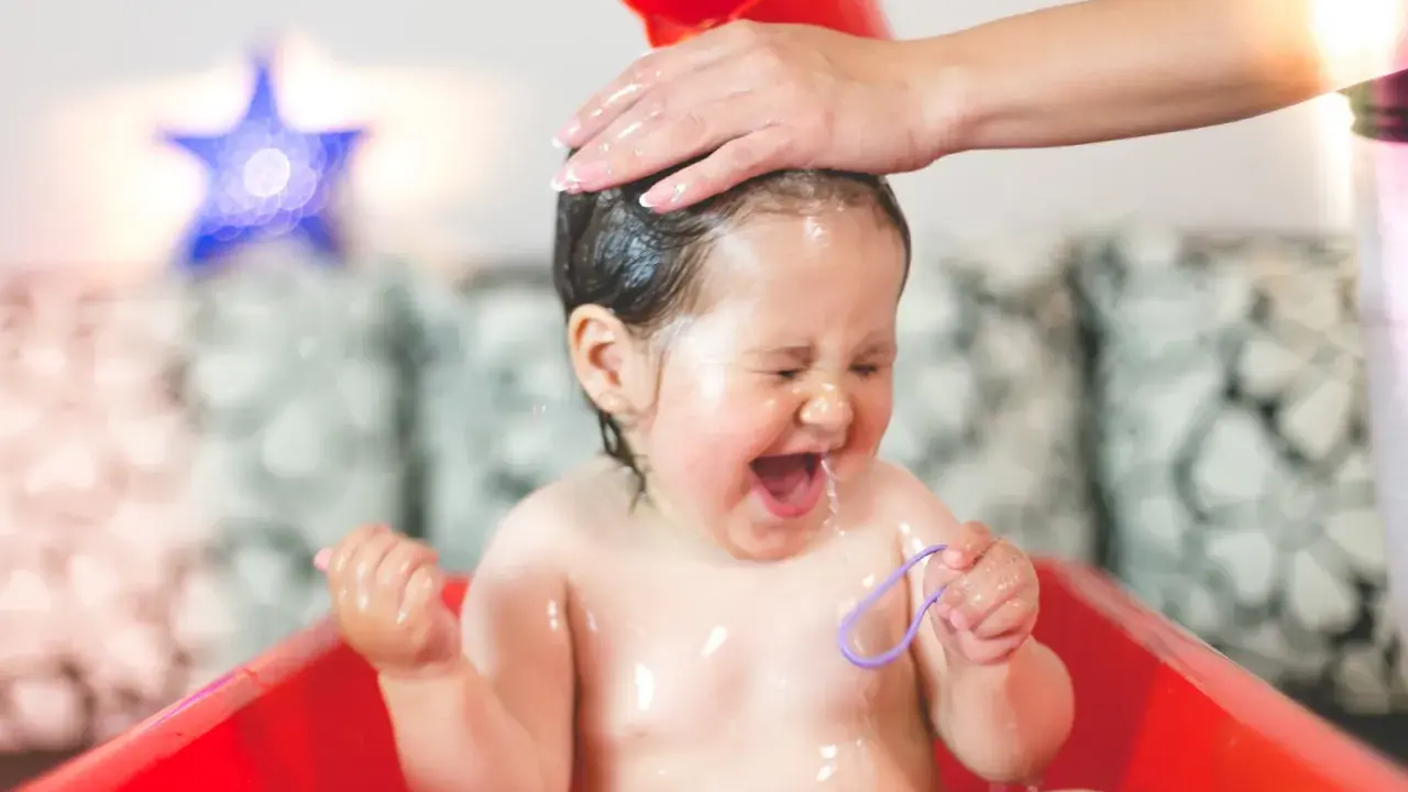 Ein Baby lacht und schreit vor Freude, während seine Haare gewaschen werden. Wasser spritzt, und es hält ein lila Band.