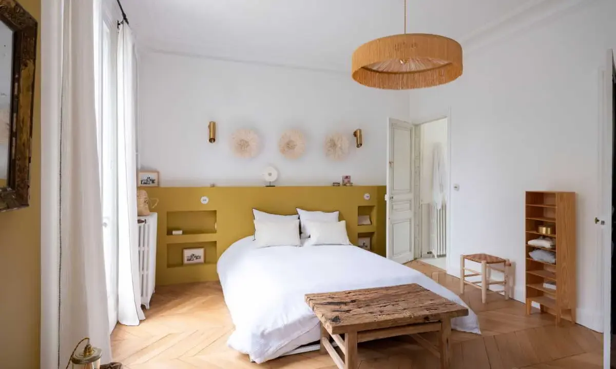 Une chambre lumineuse avec une tête de lit peinture jaune moutarde, des décorations murales en paille et un lit blanc douillet.