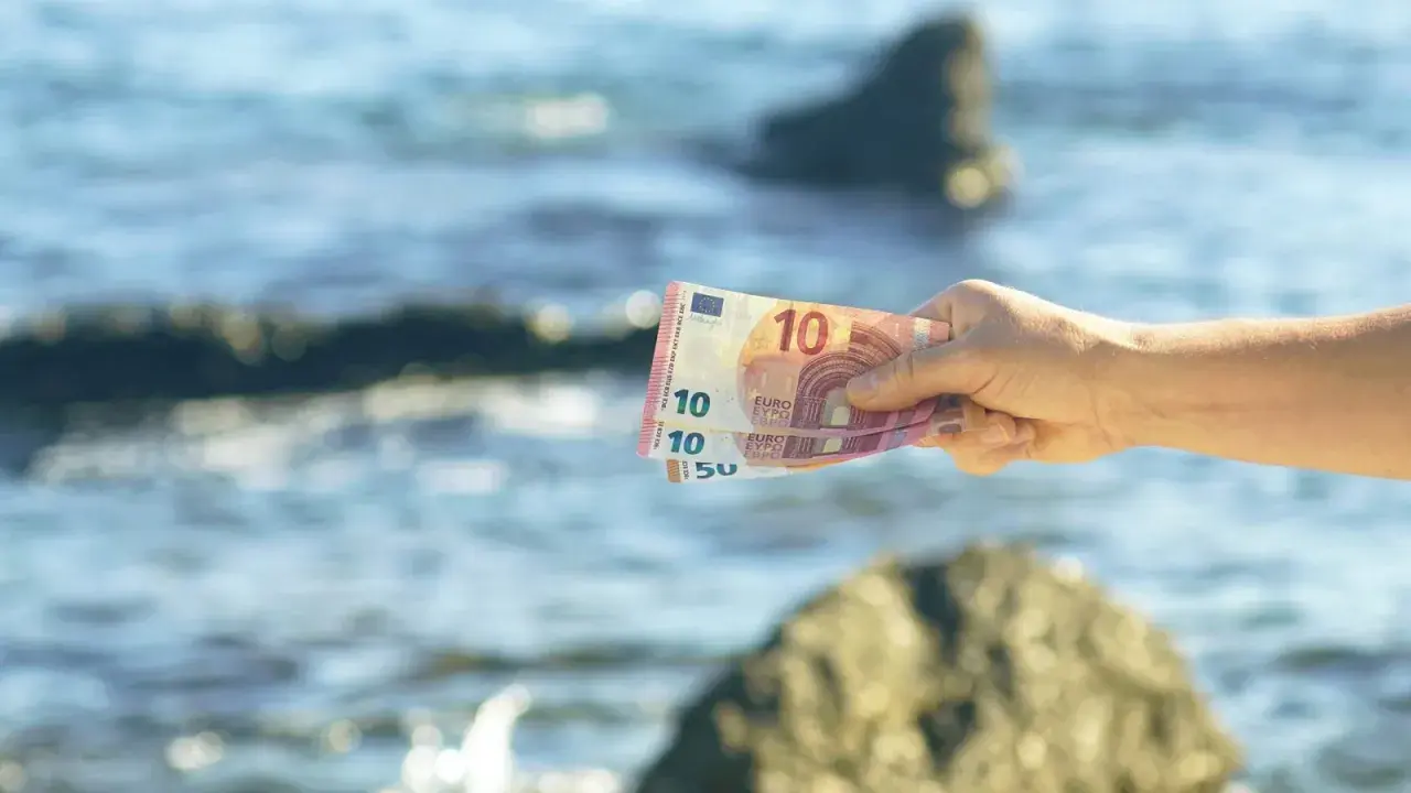Dłoń trzyma banknoty euro na tle morza. W Czarnogórze obowiązuje euro, więc te pieniądze przydadzą się na wakacjach.